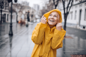 Dame steht mit gelber Regenjacke auf der nassen grauen Straße und ist gut gelaunt